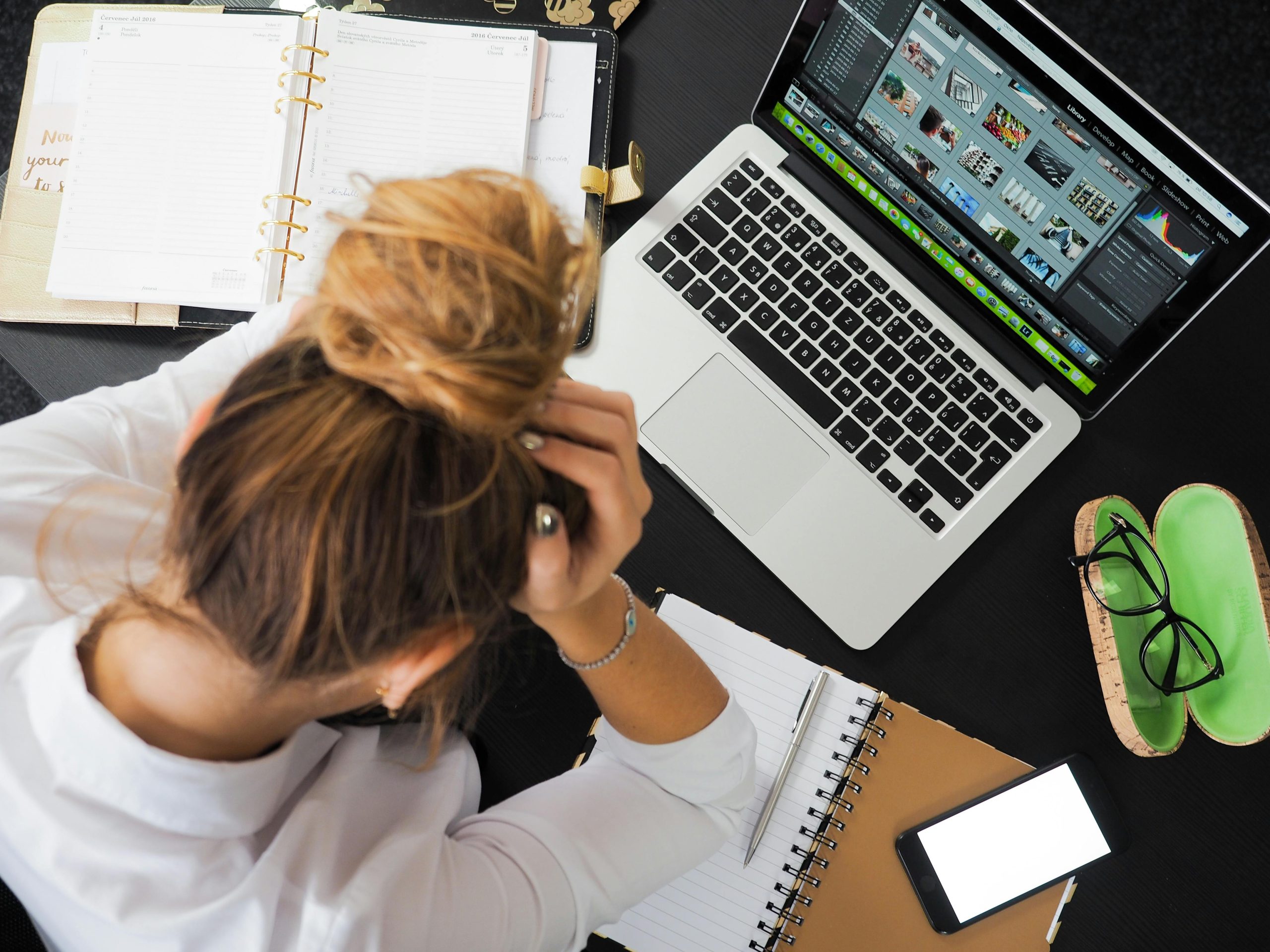 A person sat at a desk looking at a laptop, a diary and a notebook. They have one hand on their head and look as though they are conentrating. The image is taken from above.
