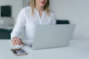 A person in an office setting looking at a laptop and usinga mouse. They are wearing a white shirt and the table is a light grey as is their laptop.