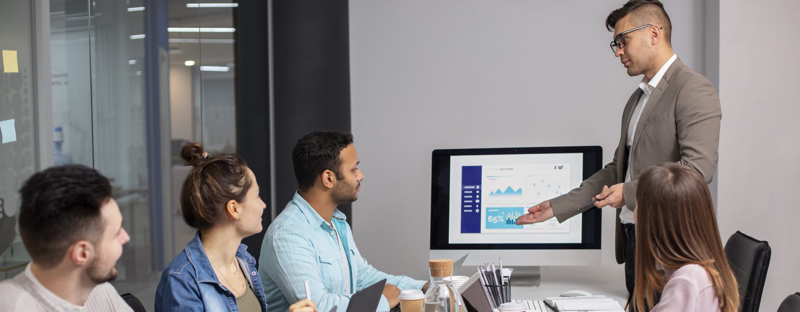 A group of 5 people in a modern looking office. They all have laptops and take away coffee cups. One of the group is standing up and making a presentation in front of a display screen. The display screen shows graphs and figures.