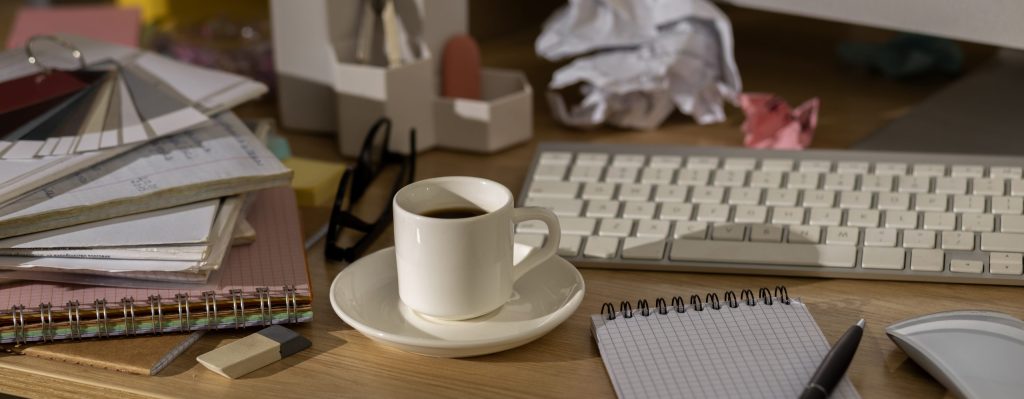 A close up of a desk with a keyboard, coffee cup and saucer, a notepad and pen and other papers and stationary.